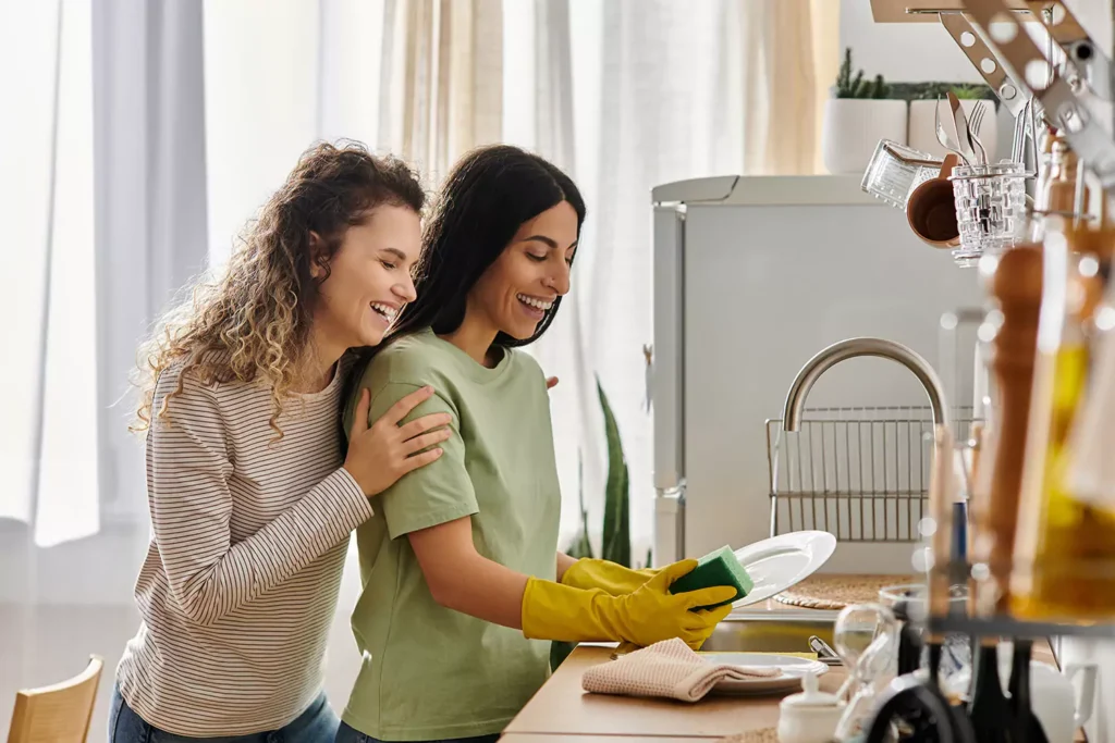 Two woman are laughing while one is washing dishes in the kitchen